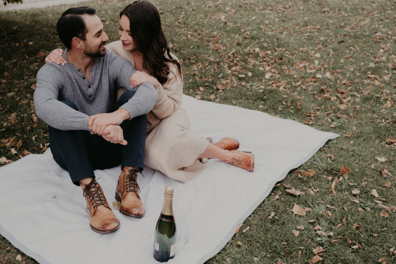 An engaged couple sitting on a blanket with a bottle of champagne.