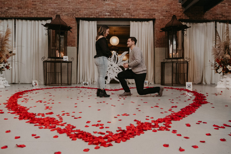 A man is kneeling down to propose to his girlfriend in a heart-shaped room, captured beautifully by Beloveful Photography.